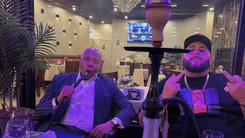 A man in a suit and pink tie smiles broadly at a podium. An American flag and a blue sign with orange text are in the background.