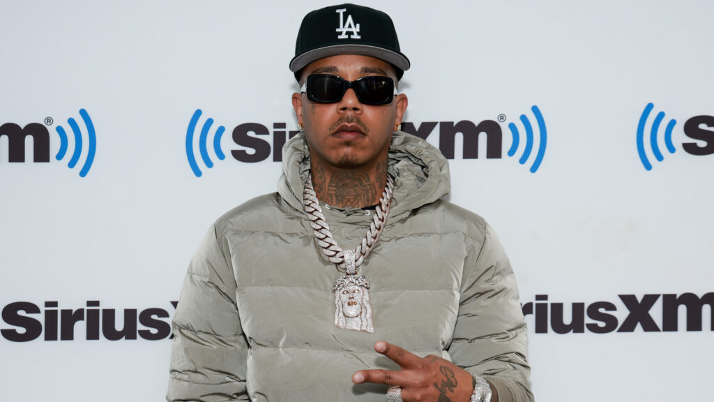 Hitmaka in sunglasses and a black LA cap, wearing a gray puffer jacket and chain, poses in front of a SiriusXM background.