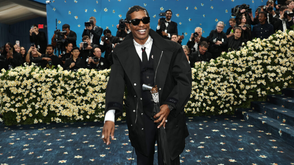 A$AP Rocky at the 2025 Met Gala, wearing a black coat and sunglasses, surrounded by photographers and a floral backdrop.