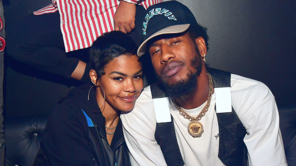 Teyana Taylor and Iman Shumpert are sitting closely together. Teyana is smiling, and Iman is wearing a cap and chain.