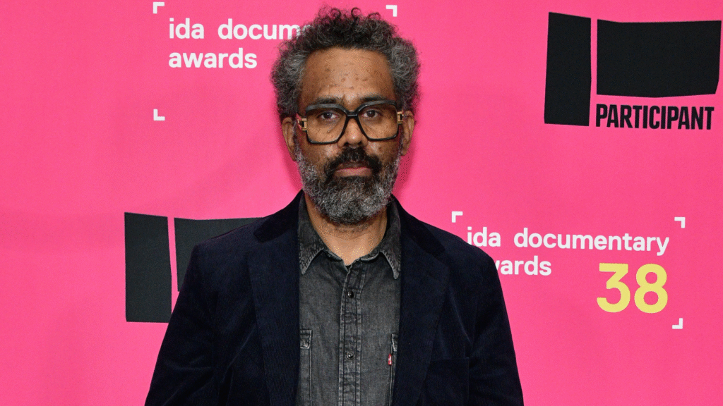 A man with glasses and a beard stands in front of a pink backdrop at the IDA Documentary Awards.