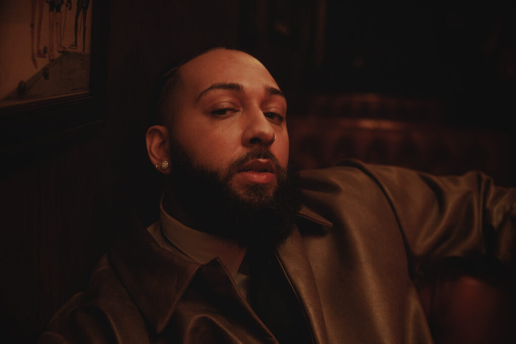 Singer Isaiah Falls with a beard and earrings, wearing a brown leather jacket, sits in a dimly lit room with a confident expression.