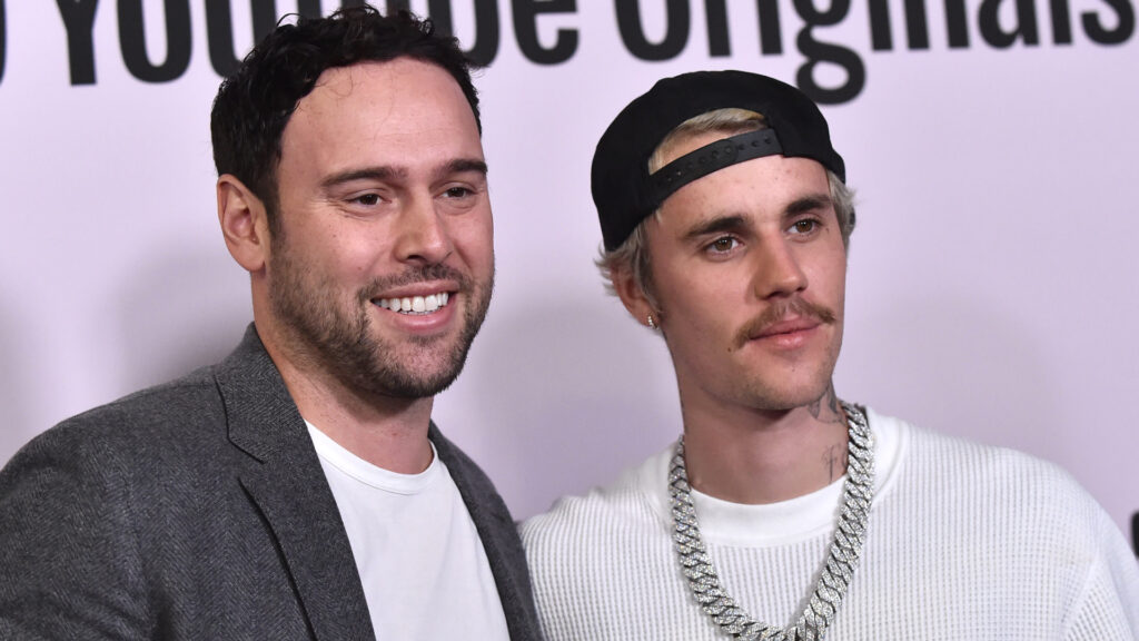 Justin Bieber Reportedly Owes Nearly $9 Million to Scoote… Scooter Braun and Justin Bieber at a YouTube Originals event. Braun in a grey blazer, Bieber in a white sweater and chain necklace.
