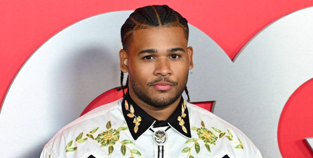A man with braided hair wearing a decorative shirt with gold and black details stands in front of a red background with a "GQ" logo.