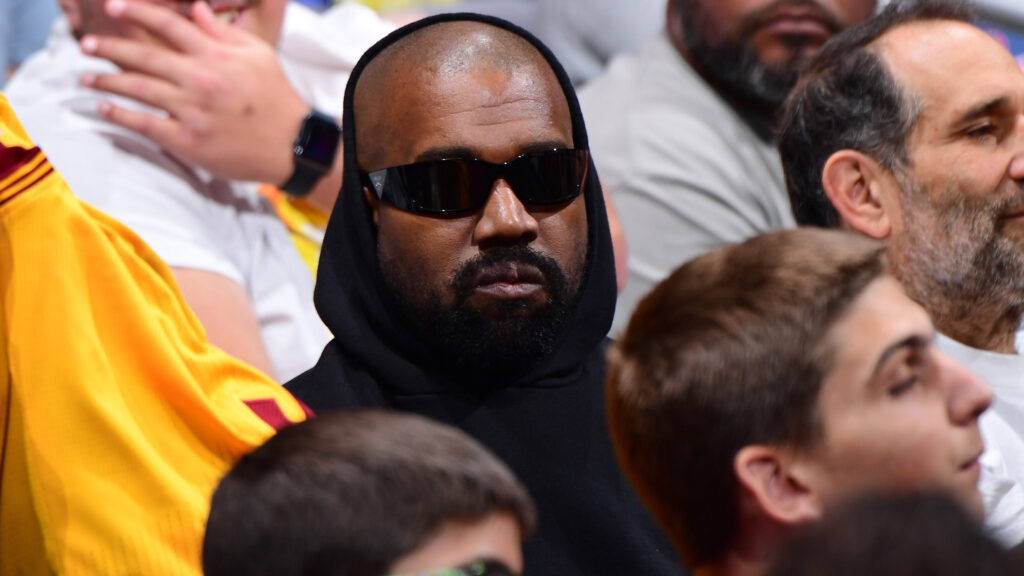 Kanye West wearing sunglasses and a black hoodie, sitting among a crowd at an event.