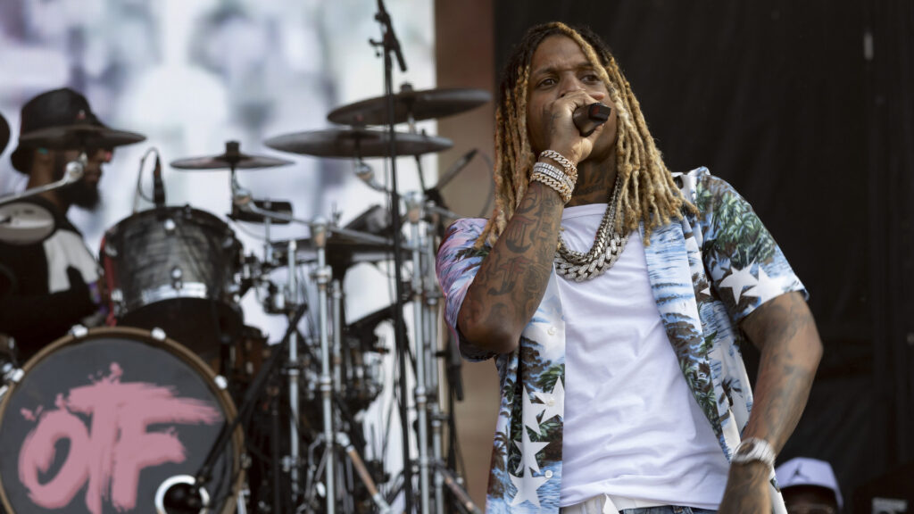 Rapper Lil Durk performing on stage, wearing a floral shirt and multiple chains, with a drummer in the background.