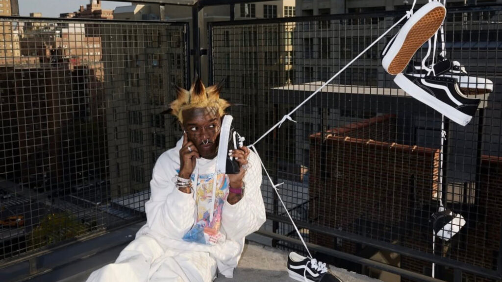 Lil Uzi Vert on Starring in New Vans Premium O… Lil Uzi vert with spiky hair sits on a rooftop, holding a sneaker to their ear like a phone. Several black-and-white sneakers hang nearby.