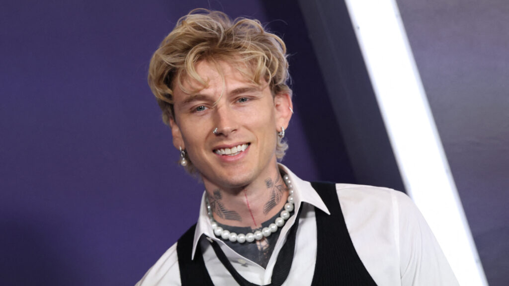 Machine Gun Kelly with blonde hair, wearing a white shirt, black vest, and pearl necklace, smiling against a purple background.