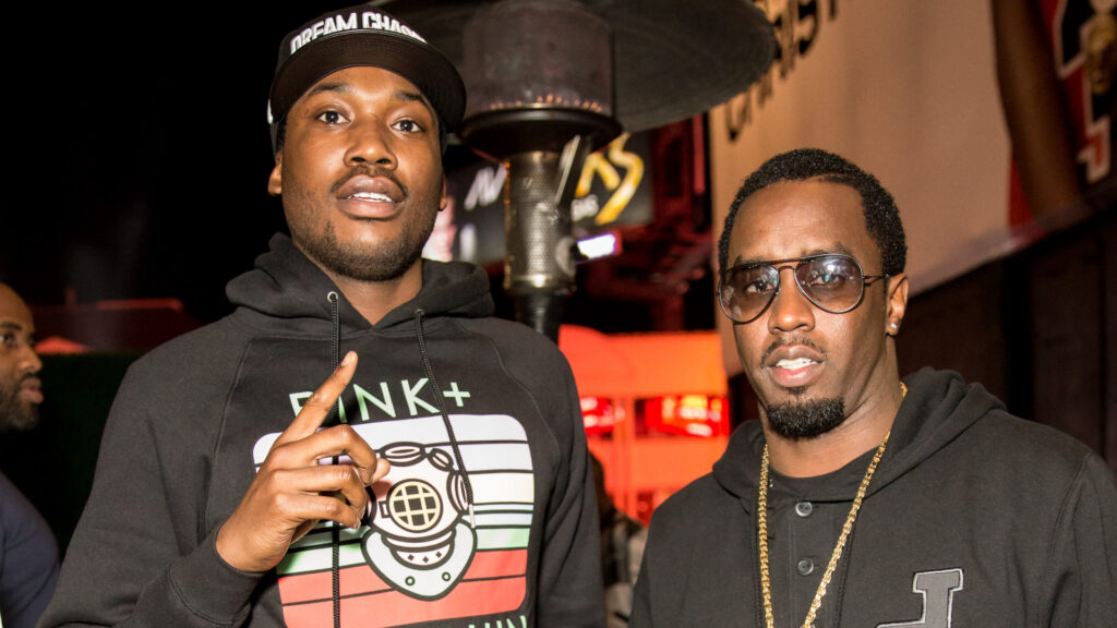 Meek Mill and Diddy wearing casual hoodies, standing side by side at an event, with a night setting in the background.