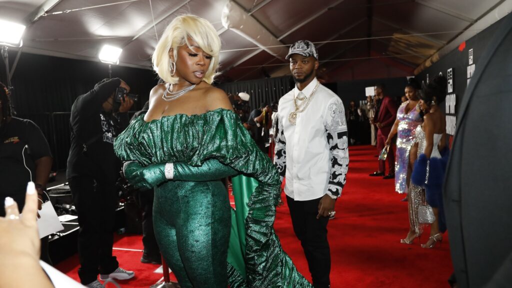 Remy Ma in a green outfit on a red carpet, with Papoose in a white shirt and cap behind her. People and photographers are in the background.