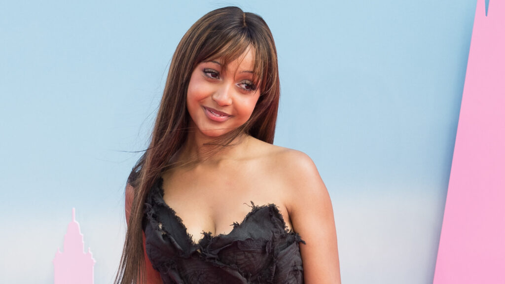 PinkPantheress in a black dress poses against a pastel background, smiling slightly with long hair framing her face.