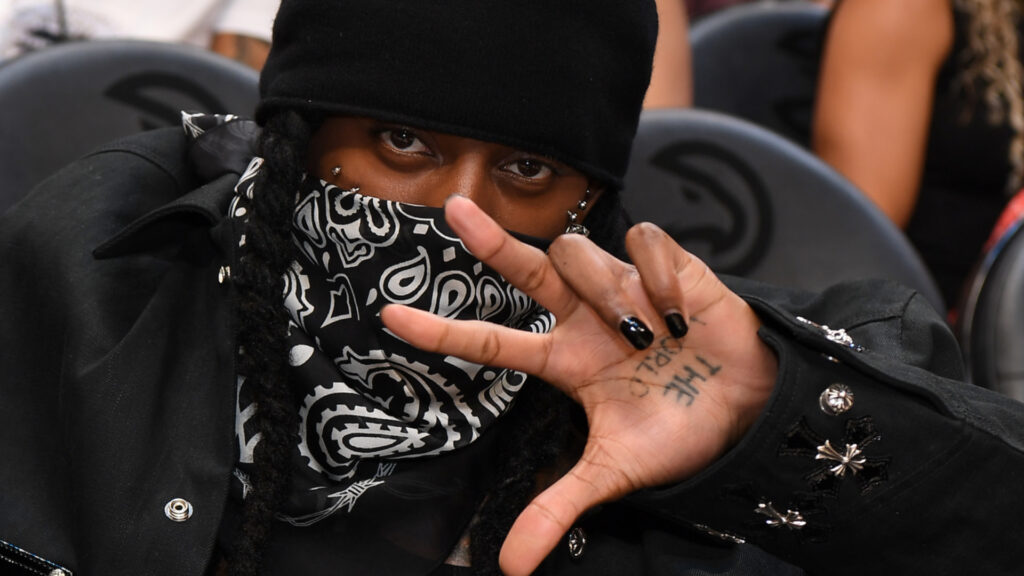 Playboi Carti in black beanie and paisley bandana covering face, showing hand with "THE" tattoo, seated indoors.