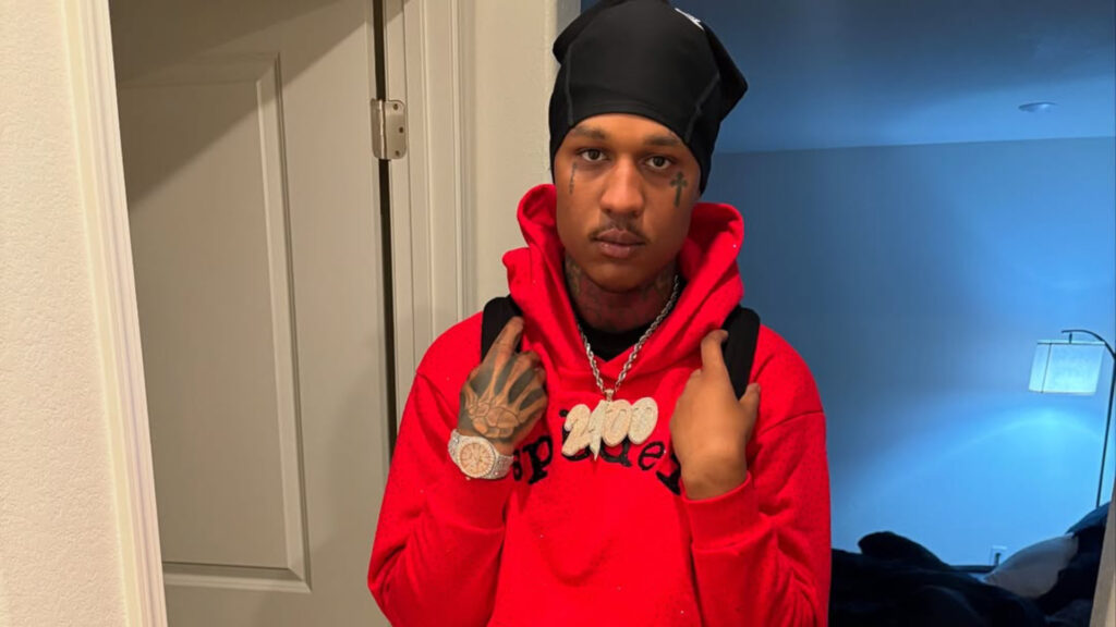 Rapper EBK Jaaybo Arrested in Arkansas on Felony Charges EBK Jaaybo in a red hoodie and black headscarf stands indoors, wearing a watch and necklace.