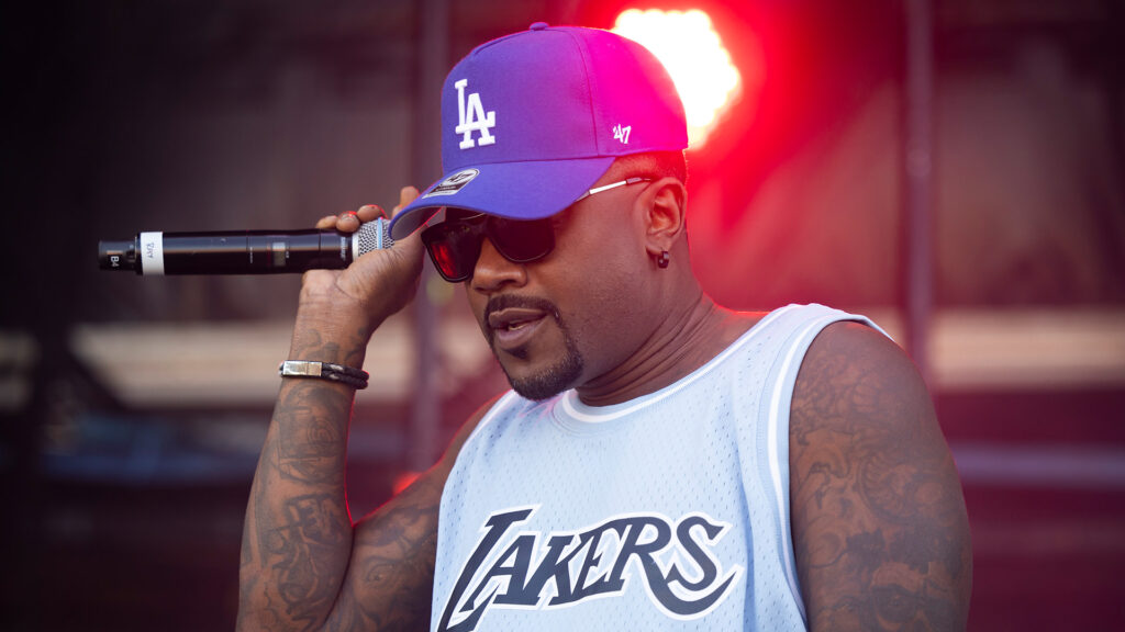 Ray J in a jersey and LA cap holds a microphone on stage, with a red light in the background.