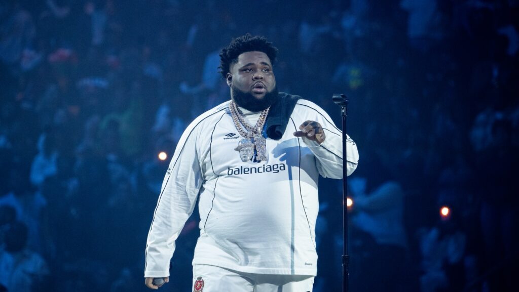 A performer in a white Balenciaga outfit sings on stage, holding a microphone. The background is filled with a cheering crowd.