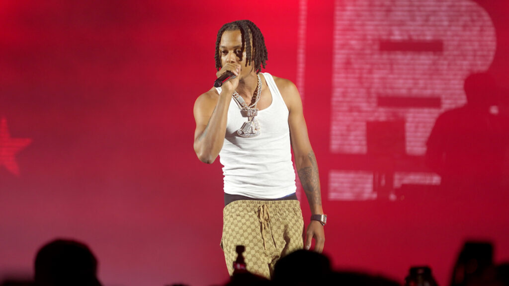 Skilla Baby in a white tank top and patterned pants performs on stage with a microphone, red lighting in the background.