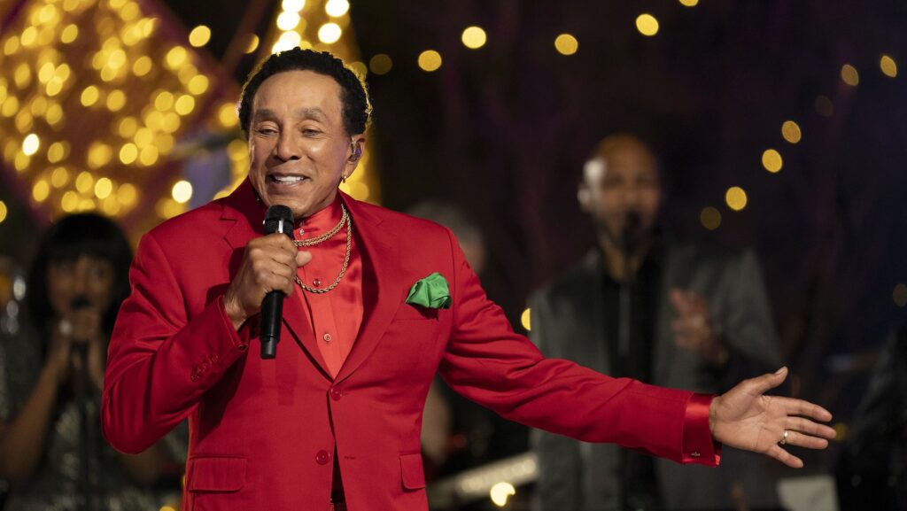Smokey Robinson, in a red suit, sings into a microphone with festive lights in the background.