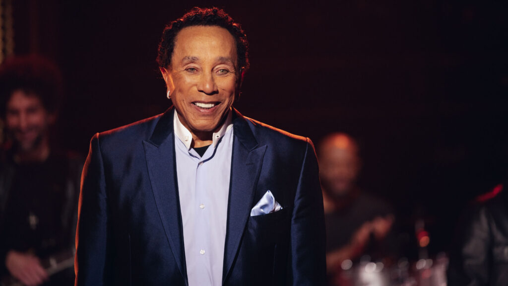 Smokey Robinson in in a blue suit and white shirt smiles on stage with musicians in the background.