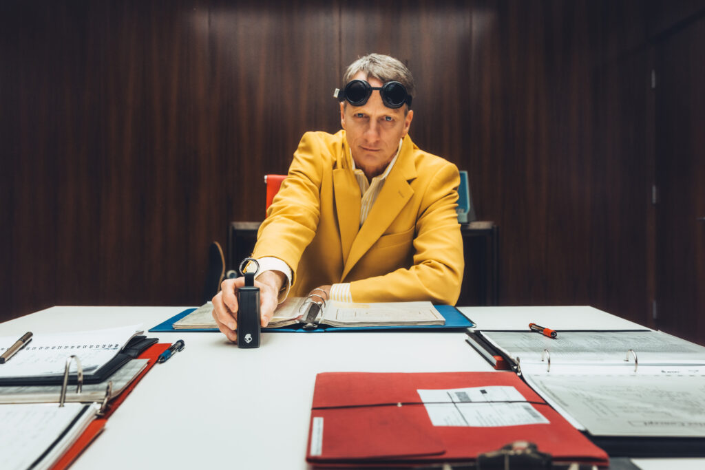 A man in a yellow suit and goggles sits at a table with binders and documents, holding a device, in a wood-paneled room.