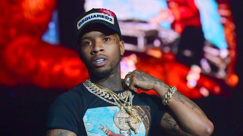 Tory Lanez wearing a "Dsquared2" cap and multiple gold chains, standing on stage with a colorful background.
