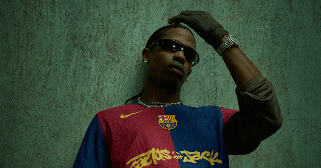 Travis Scott wearing a Barcelona jersey with "Cactus Jack" logo, sunglasses, and a glove, posing against a textured wall.