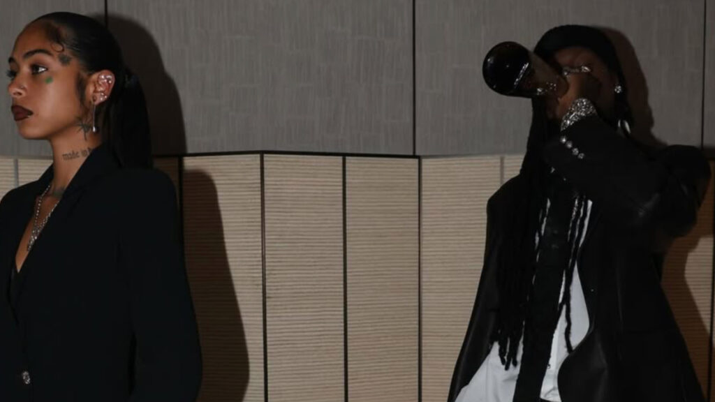 Playboi Carti wearing a beanie and tank top raises their arms on stage, with a dark background and a brick wall behind them.