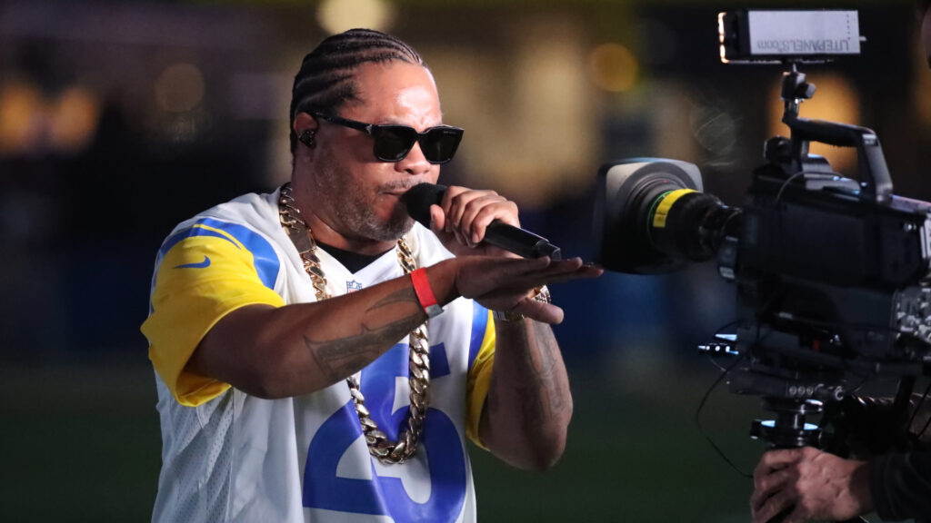 Xzibit wearing sunglasses and a sports jersey, holding a microphone, performing in front of a camera.