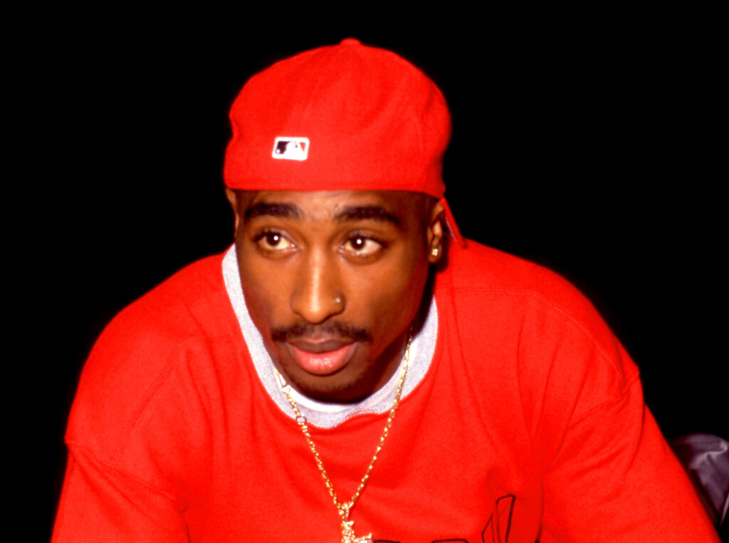 Tupac Shakur wearing a red shirt and cap, with a gold chain, against a black background.
