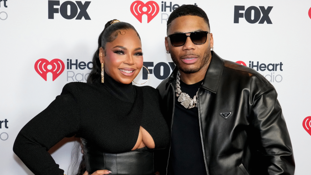 Ashanti and Nelly pose together at an iHeartRadio event. Ashanti wears a black outfit, and Nelly sports sunglasses and a leather jacket.