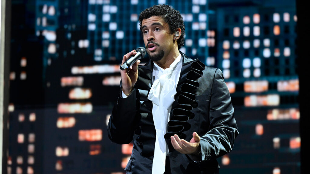 Bad Bunny in a stylish black jacket sings into a microphone on stage, with a cityscape backdrop.