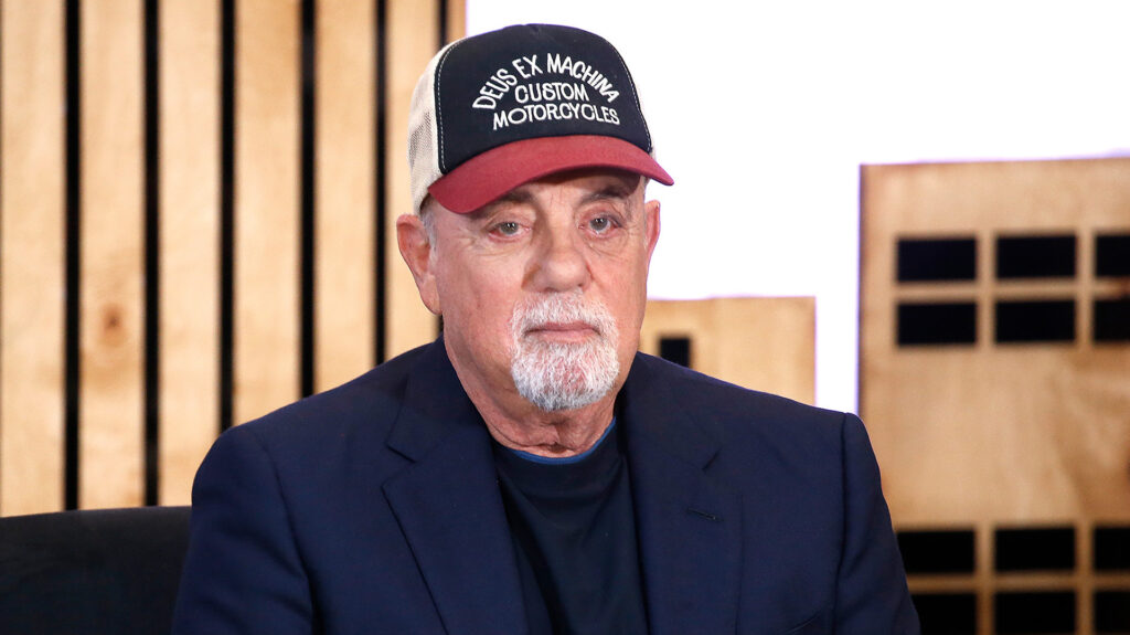 Billy Joel wearing a "Deus Ex Machina Custom Motorcycles" cap and a dark blazer, seated against a wooden backdrop.