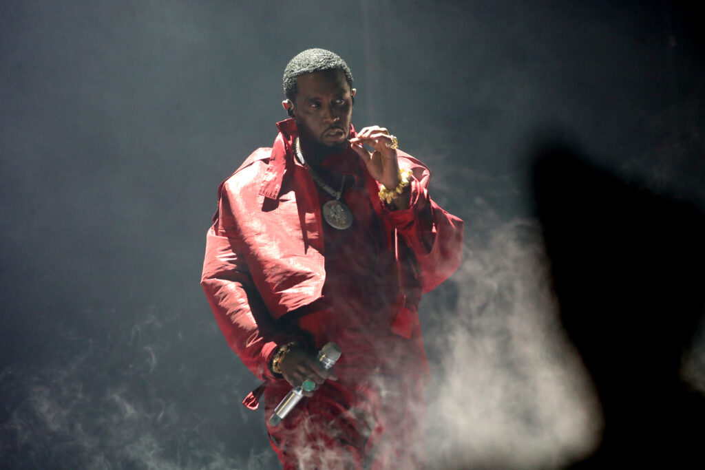 A man in a red outfit performs on stage, holding a microphone, surrounded by smoke.