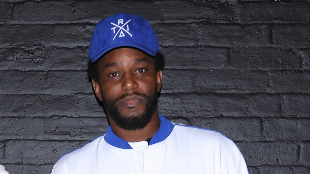 A man wearing a blue cap and white jacket stands against a black brick wall.