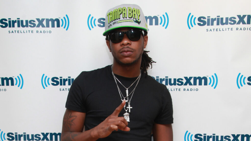 Cash Out Faces Testimony From Sex Trafficking Expert in C… Rapper Cash Out wearing a "Tampa Bay" cap, sunglasses, and necklaces, posing in front of a backdrop.