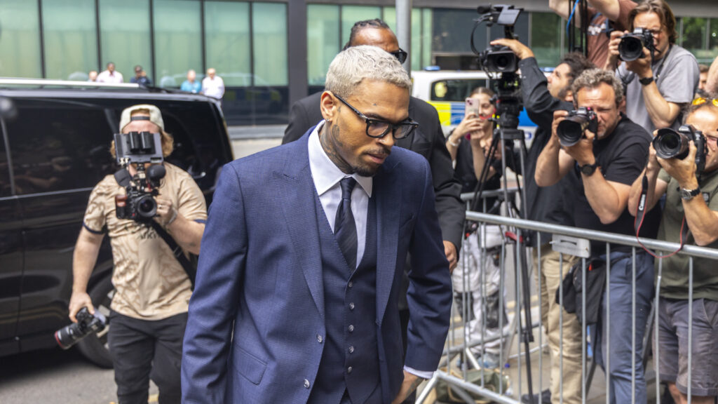 Chris Brown in a blue suit with glasses walks past photographers outside a building.