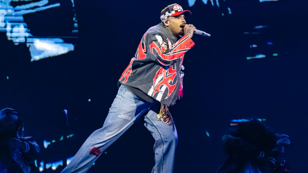 Chris Brown on stage wearing a colorful jacket and jeans, holding a microphone, with a dynamic pose against a dark background.