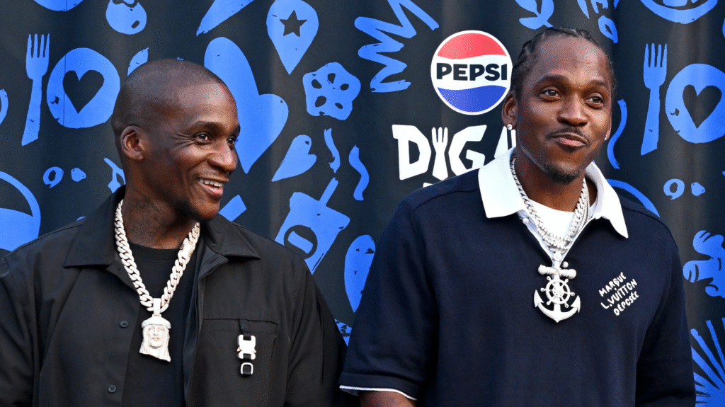 Two men stand in front of a Pepsi backdrop, both smiling. They wear black shirts and large chains.