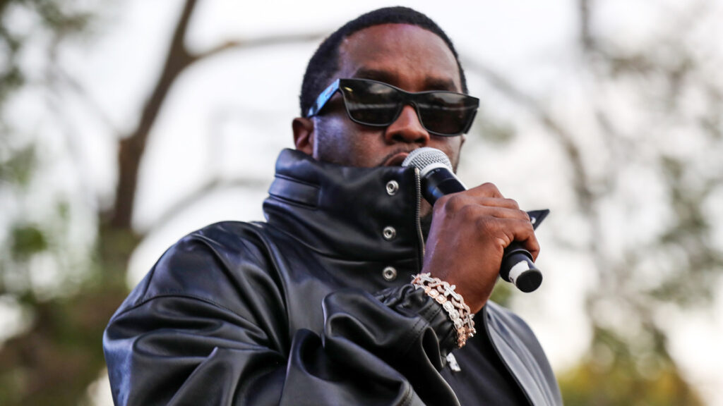Diddy in a black leather jacket and sunglasses holds a microphone, performing outdoors.