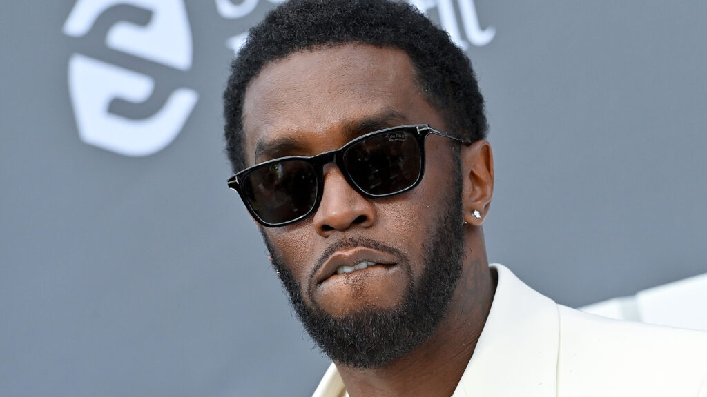 Sean "Diddy" Combs wearing sunglasses and a white suit, with a serious expression at an event.