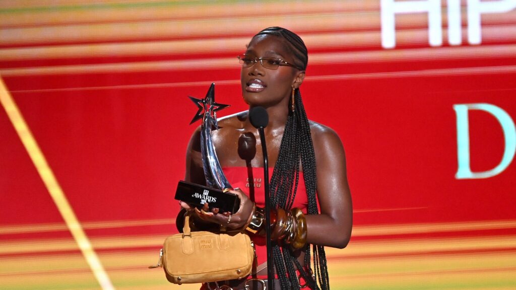 A woman with long braids, wearing glasses and a red dress, holds a trophy on stage at an awards event.