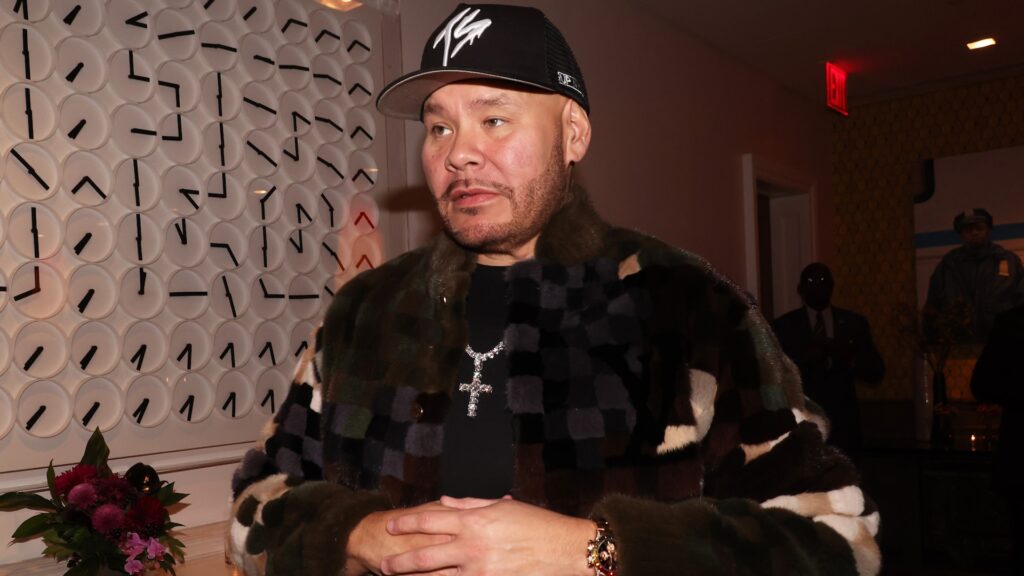 Fat Joe wearing a patterned jacket and a cap, standing indoors with a decorative wall behind him.