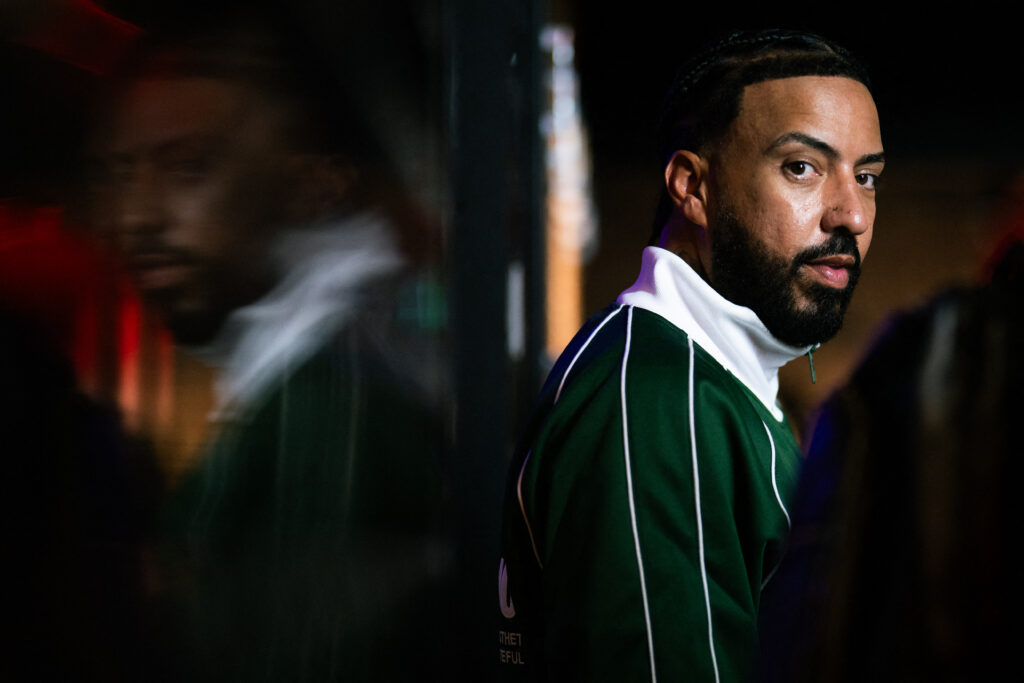 French Montana wears a green jacket with white stripes stands near a reflective surface, looking sideways. The lighting is dim and moody.