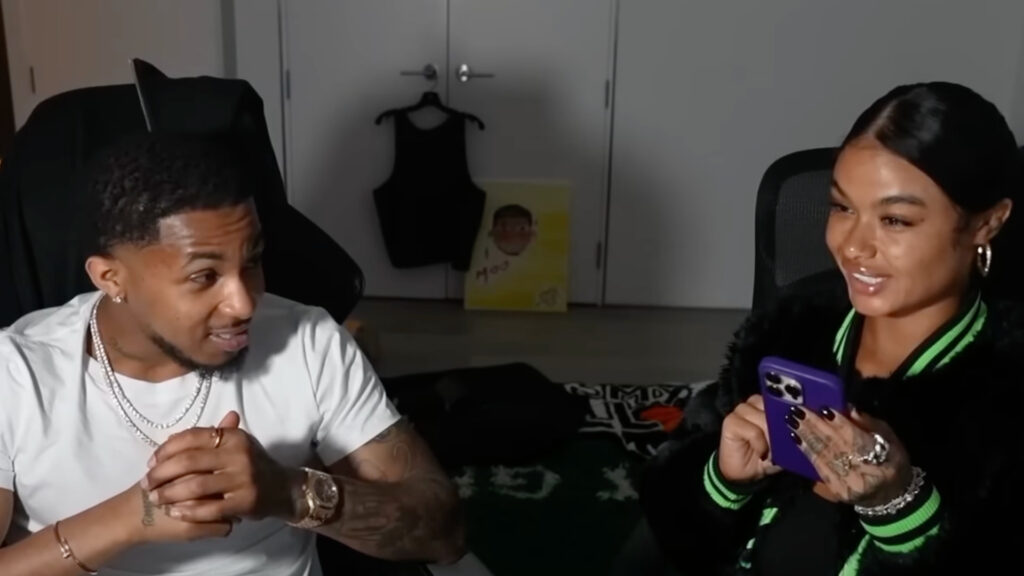 India Love wears a purple outfit with gold jewelry, and DDG wears a black shirt with layered necklaces.