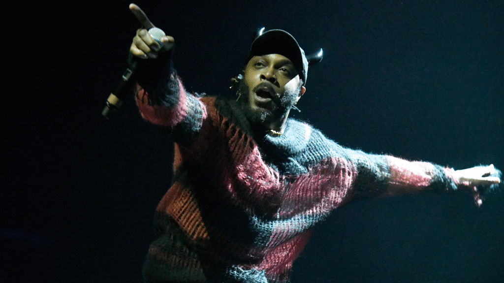 A performer on stage wearing a dark cap with small horns, a red and black sweater, holding a microphone and pointing outward.