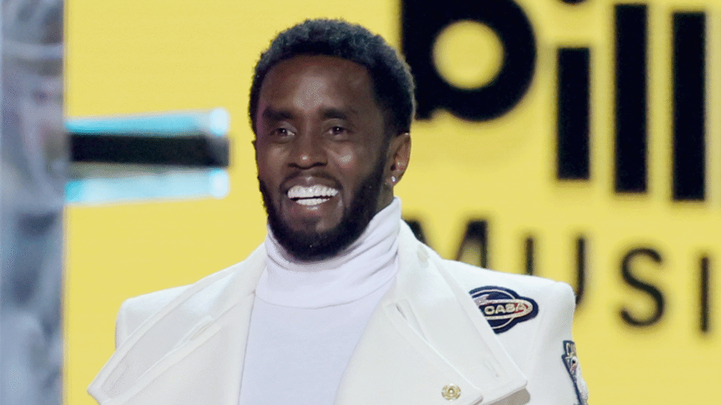 Sean "Diddy" Combs smiling, wearing a white turtleneck and jacket, at an event with "Billboard Music" signage in the background.