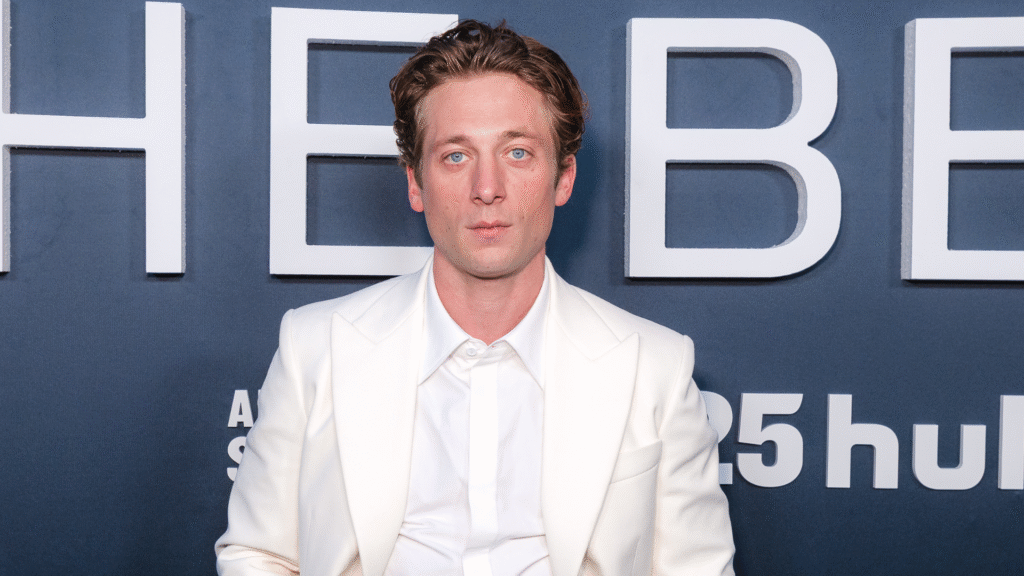 A man in a white suit poses in front of a backdrop with text.