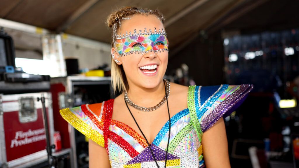 JoJo Siwa in a colorful, sparkling costume and mask, smiling backstage.