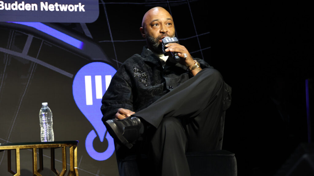 Joe Budden sitting on stage, speaking into a microphone, with a water bottle on a table nearby.