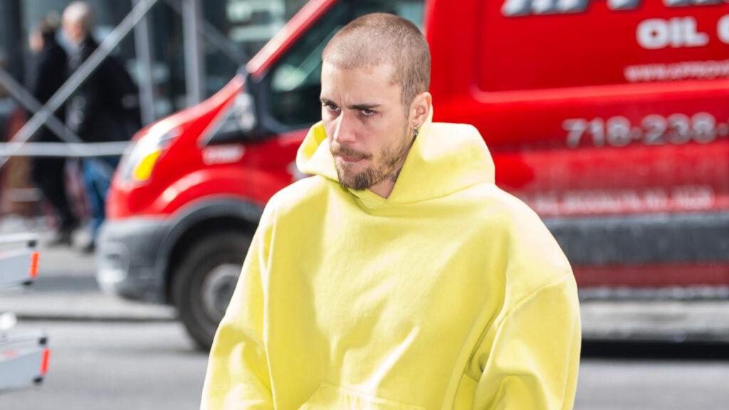 Justin Bieber in a yellow hoodie walks on a street with a red van in the background.