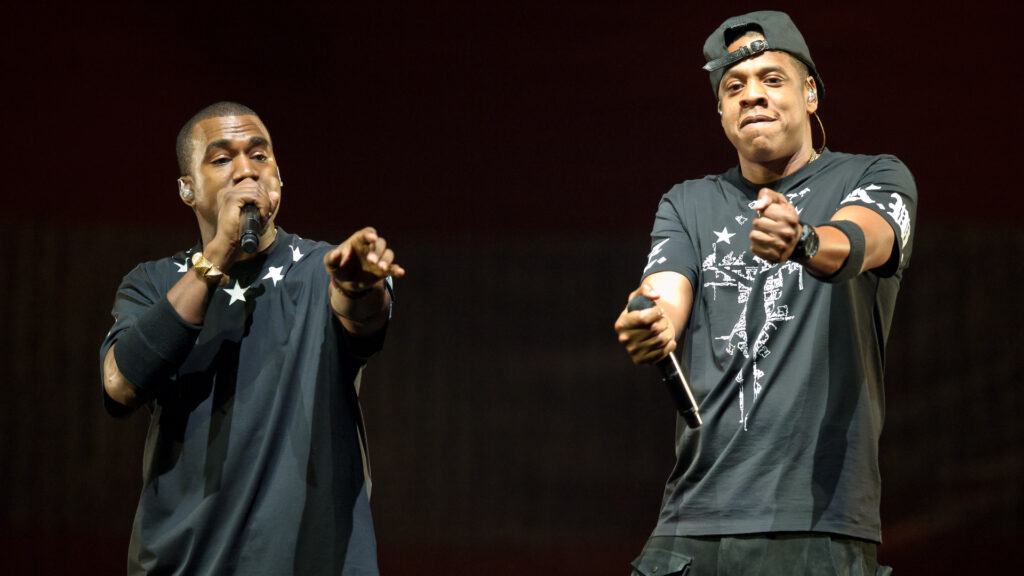 Kanye West and Jay-Z performing on stage, both wearing dark shirts, holding microphones, and engaging with the audience.